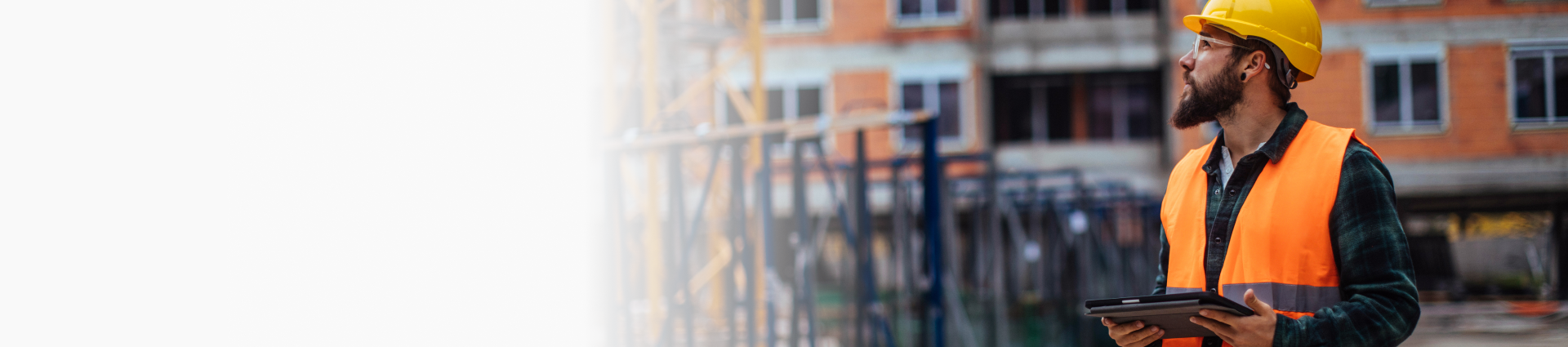 Construction worker in high-visibility clothing and a hard hat holds a tablet at a building site.