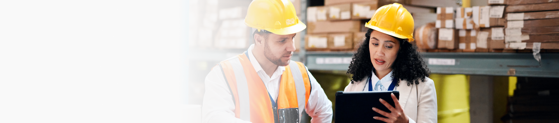 Two workers in a warehouse, one holding a tablet. Shelves stocked with items in the background.